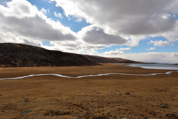 A narrow winding river crossing a flat yellowed steppe flows into a large frozen lake on an autumn cloudy day.