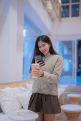 Young asian woman is using her smartphone and drinking takeaway coffee inside a modern cafe, enjoying a relaxing moment with technology and beverage