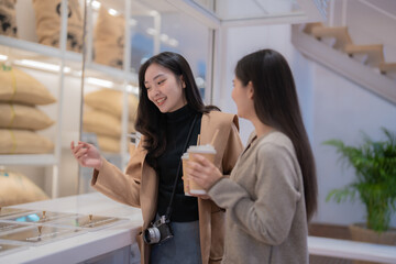 Two young women, holding takeaway coffee cups, are selecting coffee beans in a modern coffee shop, savoring their leisure time together while chatting and enjoying each other's company