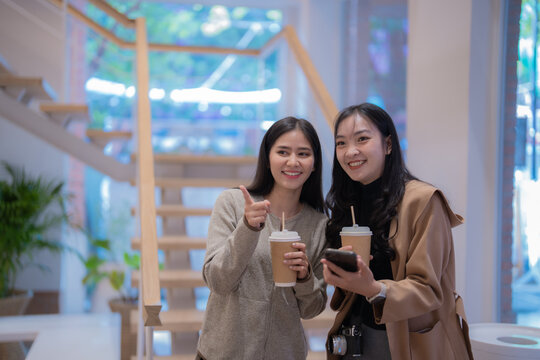 Two young Asian women enjoying takeaway coffee, smiling and giving thumbs up while using their smartphones indoors in a modern building filled with stairs and greenery