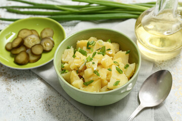 Bowl of tasty potato salad with green onion, pickled cucumber slices and oil on light grunge background