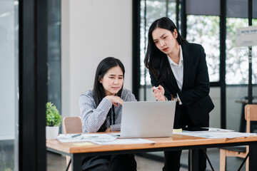 Two businesswomen in a modern office setting, collaborating on a project using a laptop and documents.