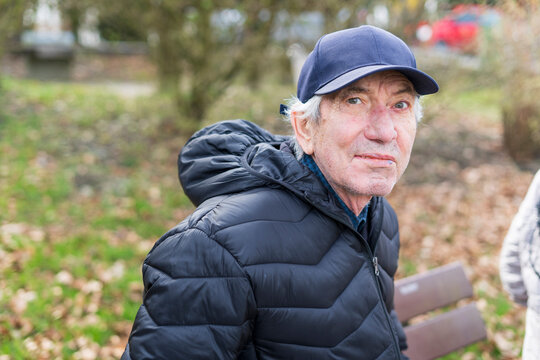 An elderly man in a jacket and hat sits alone on a bench in a park. The concept of loneliness in old age