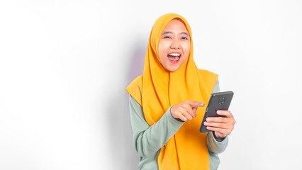 Excited Asian muslim woman wearing yellow hijab holding mobile phone and pointing at it while...