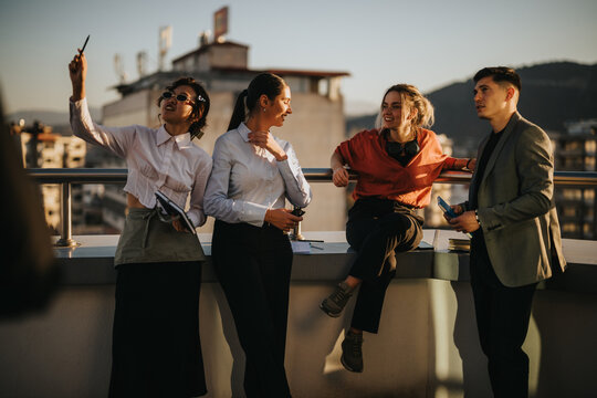 A group of multiracial business colleagues relaxes on a rooftop balcony, celebrating a successful day as the sun sets over the cityscape, showcasing camaraderie and diversity in a professional setting - Powered by Adobe