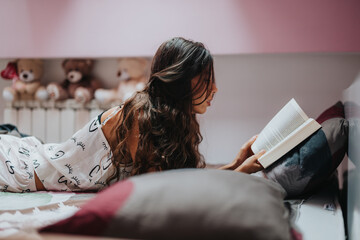 A young woman with long hair reads a book while relaxing in a comfortable bedroom, surrounded by...