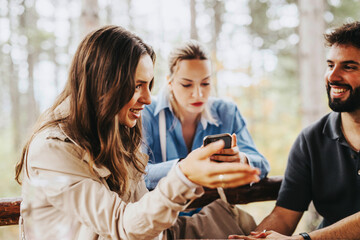 A group of friends engaging in lively conversation outdoors. They are smiling and using a smart phone, surrounded by a serene forest background, creating a warm and joyful atmosphere.