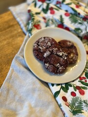Chocolate red velvet crinkle Christmas cookies served on a plain stoneware plate, surrounded by a festive tablecloth featuring holiday patterns, evoking warmth and celebration.