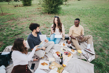 A diverse group of friends sitting on a blanket sharing food and drinks during a picnic in a green...