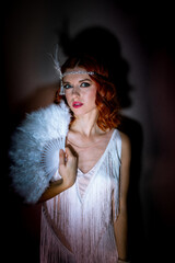 Retro 1920s style woman with red hair wearing a light fringed dress. He is holding a white feather...