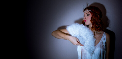 Retro 1920s style woman with red hair wearing a light fringed dress. He is holding a white feather...