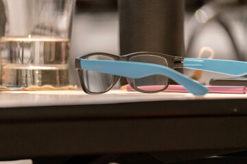 Close-up of eyeglasses with bright blue temples resting on a desk. Surrounding of office supplies, workspace ambiance.
