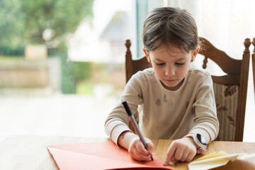 Naklejka premium Little cute girl doing origami sitting at the table with a big window in the background. Children's education and creativity concept