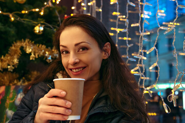 Beautiful tothy smiling woman drinking coffee in city cafe near the window, looking in camera with near Merry christmas decoration, bright lights anf sits near xmas tree. Closeup