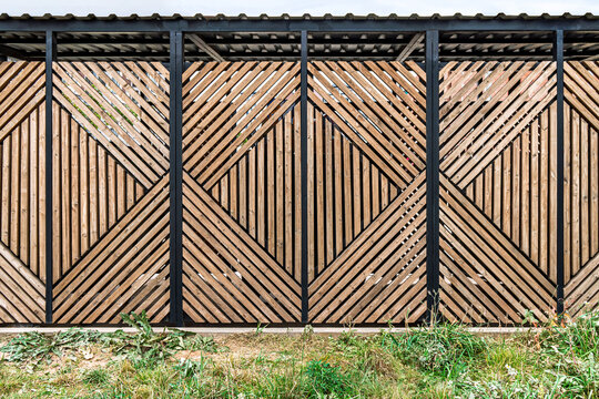 Geometric wooden fence with black metal frame, intricate angled patterns, outdoor setting, overgrown grass, design concept
