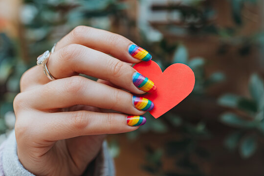 Close-up of a hand with rainbow-colored nails holding a heart, symbolizing pride and acceptance