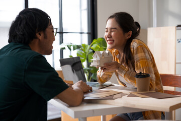 Asian couple sitting in their home, looking at house plans and models, and discussing plans for their dream home. Working together, planning, and financial aspirations.