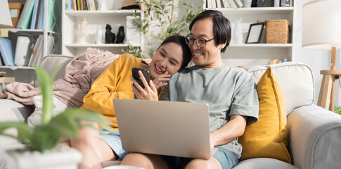 Asian woman showing her smartphone with interesting content, online shopping videos, to her husband. They laugh and enjoy the moment together while husband works on his laptop in their living room.