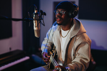 A young artist plays an electric guitar and sings into a microphone in a studio, creating music....