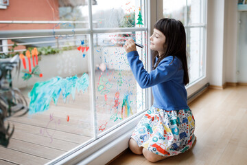 Cute little girl drawing merry christmas wishes on window