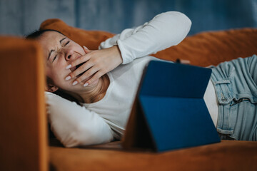A young woman yawns widely, relaxing on a comfortable couch while watching a tablet. The scene captures a moment of relaxation and fatigue in a casual home setting.