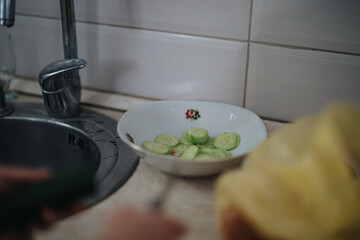 A bowl of fresh cucumber slices is placed beside a kitchen sink, capturing a moment of home cooking preparation. The setting evokes a sense of simple, everyday kitchen life.