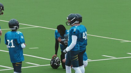 Diverse American Football Players in Blue Jerseys Having a Time Out. Resting on a Field, Talking with Teammates. Black Footballer is Sitting with His Helmet Off and Having a Strategic Conversation - Powered by Adobe