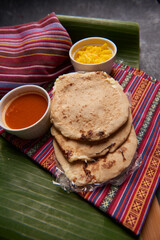Pupusas with curtido and sauce served on a tamale leaf atop a wooden surface, highlighting traditional Central American flavors.