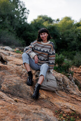 Naklejka premium young woman in a stylish sweater enjoys a day in the mountains, sitting on the rocks, with a picturesque landscape opening behind her. hiking and walking.