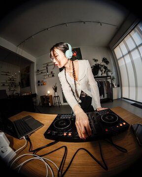 smiling young woman DJ wearing mint headphones mixes music on a DJ controller in a cozy home studio with shelves, houseplants, and decorative elements in the background.