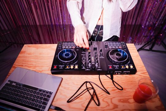 DJ's hands are adjusting the controls on a professional mixer placed on a wooden table, with a laptop and drink nearby, illuminated by colorful pink and blue lighting.