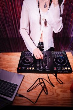 DJ's hands are adjusting the controls on a professional mixer placed on a wooden table, with a laptop and drink nearby, illuminated by colorful pink and blue lighting.