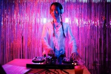 DJ wearing a white blouse and headphones performs on a professional mixer, standing against a vivid backdrop of pink and blue streamers, enhancing the club atmosphere.