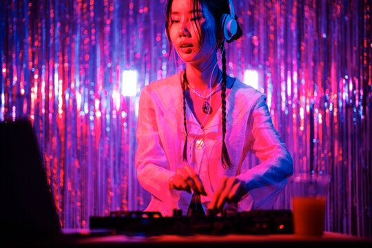 DJ wearing a white blouse and headphones performs on a professional mixer, standing against a vivid backdrop of pink and blue streamers, enhancing the club atmosphere.
