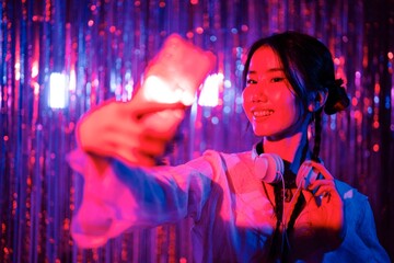 DJ with headphones and a bright smile takes a selfie with a smartphone, standing against a vivid backdrop of pink and blue streamers in a colorful and energetic club setting.