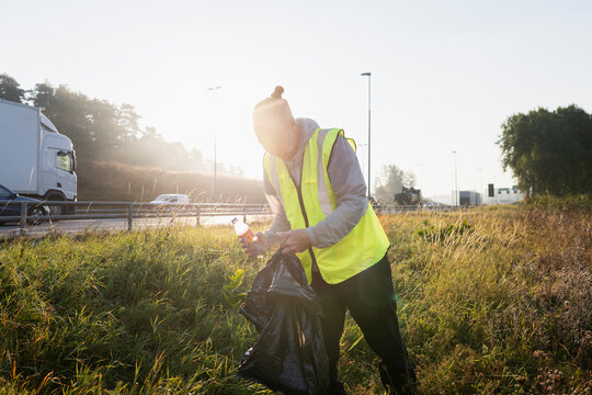 Male sanitation worker collecting garbage while standing on grass near road at sunny day