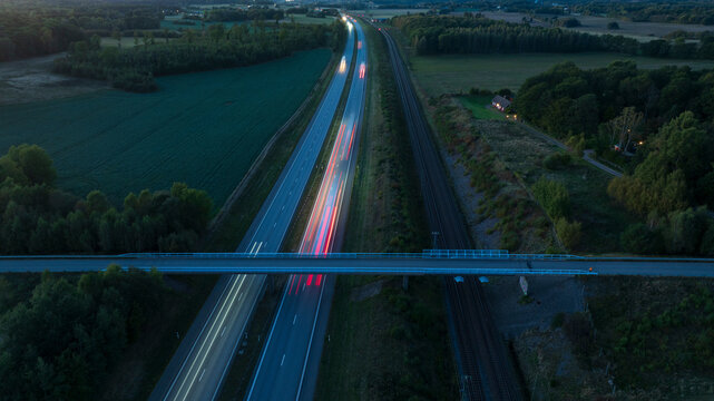 Drone view of bridge above two lane illuminated highway amidst agricultural field at dusk