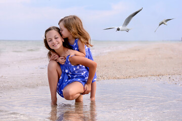 Fototapeta premium Family photo of sisters. Girls are walking on the beach near the sea, looking for a shell, smiling, hugging, they are happy and joyful. Dressed in identical dresses. Blurred background.