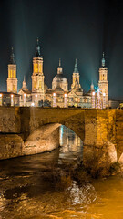 Christmas lights envelop the Basilica del Pilar, while the stone bridge and the reflection of the Ebro River create a dreamy night scene in the city of Zaragoza.