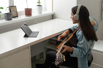 Latin girl with headphones listening to her online music lessons. Artistic kid playing the acoustic guitar and learning the chords