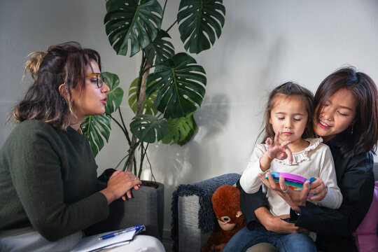 Psychologist observing little girl playing fidget toy with her mother during therapy session