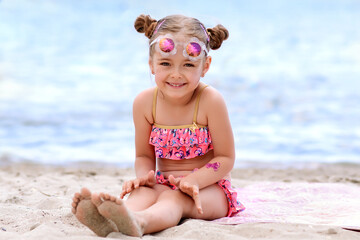 A little very cheerful girl sits on the sand on the seashore in sunglasses in the summer. The girl has an inflatable ball and an inflatable circle. Family vacation on the seashore or ocean.