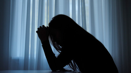 Silhouette of woman suffering mental health issues, sitting against window, depressed expression,...