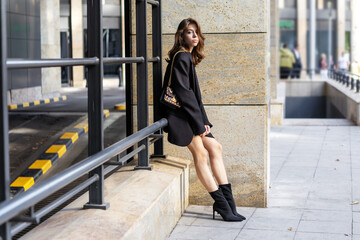 Young brunette woman stands, poses in urban city, beautiful model wearing elegant outfit oversize black blazer and trendy Heeled boots. Modern high fashion, street style on Urban background