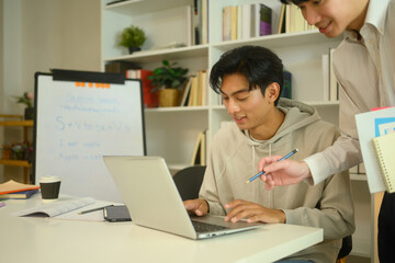 Asian male student in a hoodie working on a laptop, guided by a tutor at home