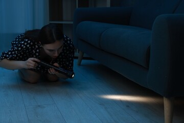 Fear of darkness. Scared young woman looking under couch with flashlight indoors at night, space...
