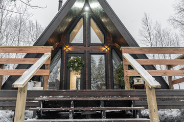 small A-frame cabin with natural wood siding, floor-to-ceiling glass panels at front. narrow wooden deck wrap around house. wooden house in snow-covered coniferous forest. Glamping in winter.