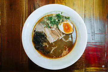 Japanese Ramen with dark broth and topped with slices of tender pork, soft-boiled egg, seaweed, green onions, and sesame seeds. Flat lay with wooden table.
