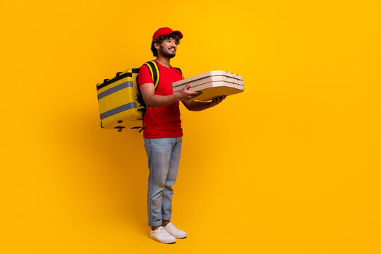 Young courier wearing red uniform holding pizza boxes while standing with yellow thermal bag against yellow background - Powered by Adobe