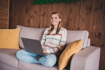 Young woman with blond hair relaxing at home in casual style with a laptop, surrounded by cozy cushions in a modern living room